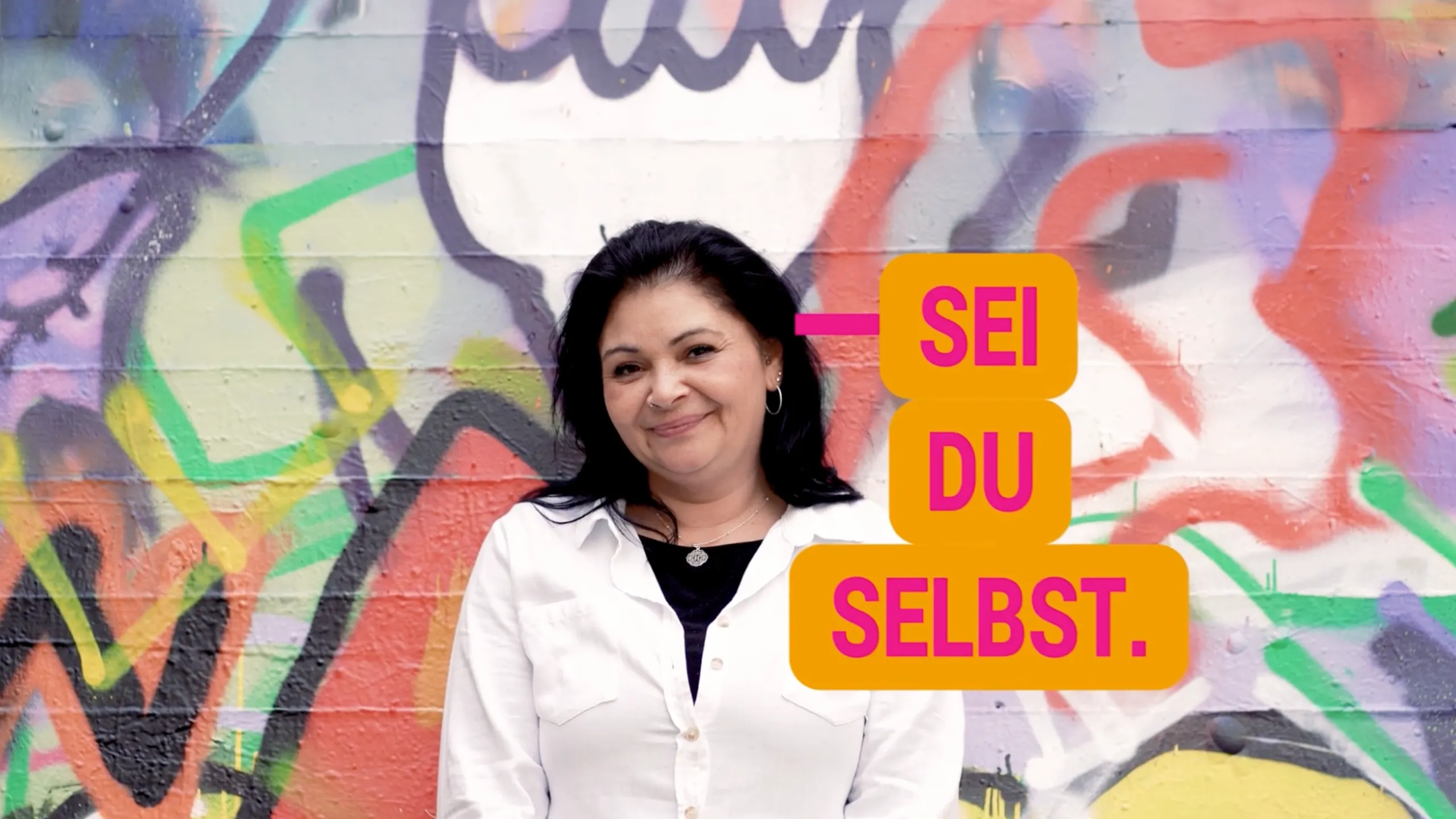 Frau vor bunter Graffiti-Wand mit Text „Sei du selbst“ – Recruiting-Kampagne Pflege zu Hause
