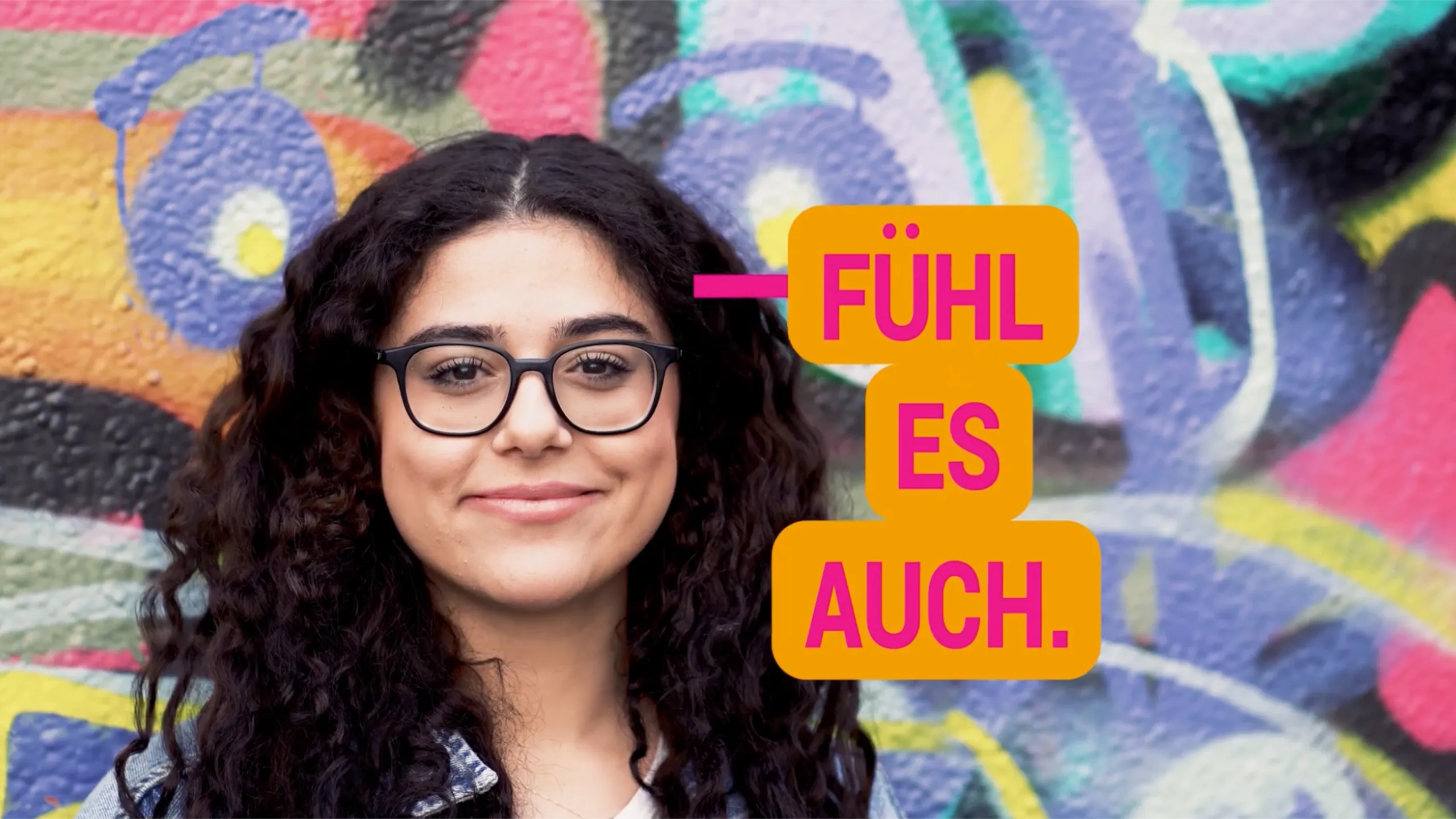 Junge Frau mit lockigem Haar vor Graffiti-Wand mit Text „Fühl es auch“ – Kampagne Pflegeberufe