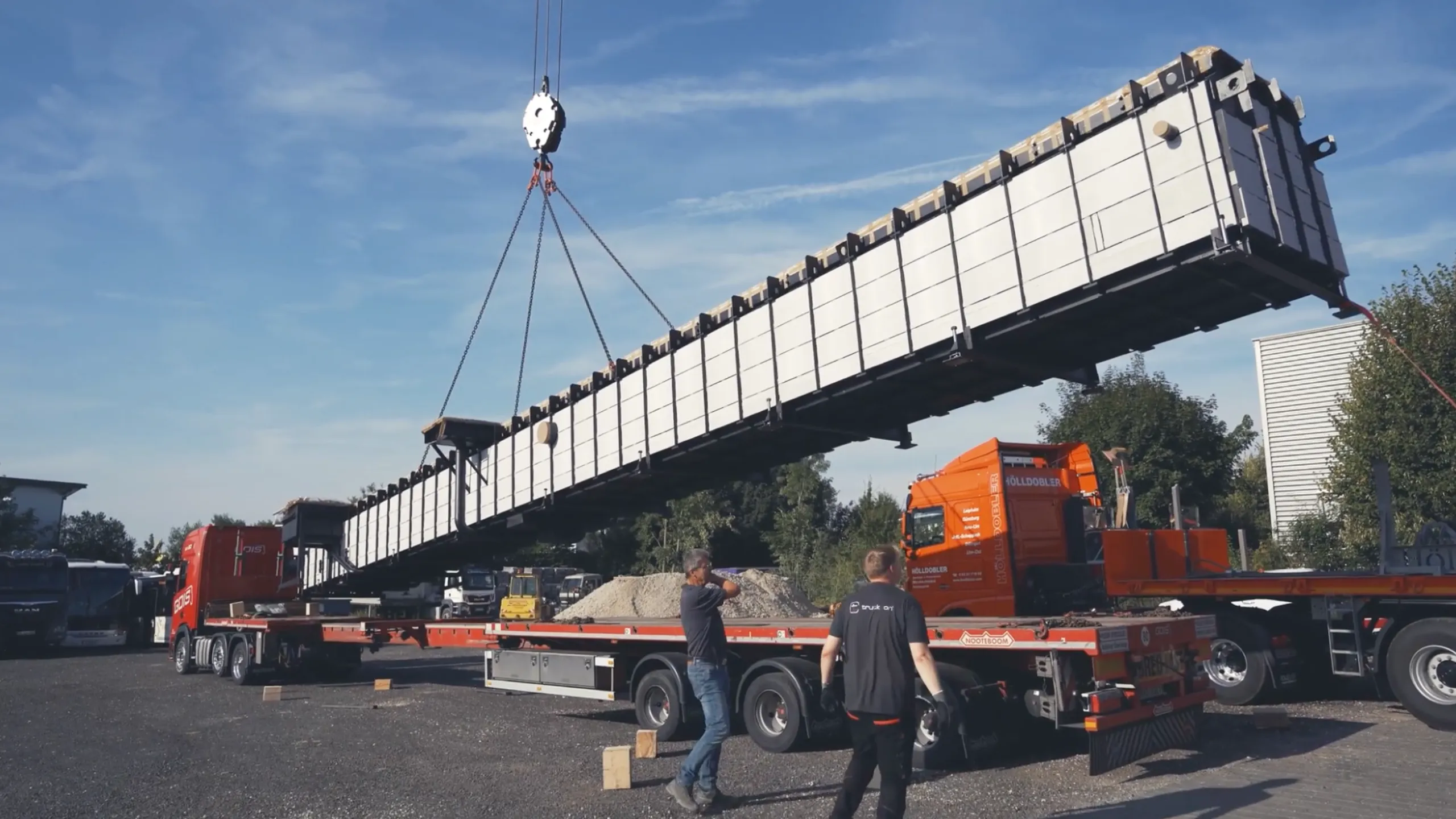 Kran hebt eine Stahlfertiggrube auf einen Tieflader während der Montage auf einem Werkstattgelände – truckcon Einbauprozess